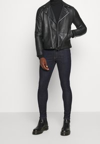 Chaqueta de motociclista de cuero negro con cremalleras plateadas, cuello alto negro ajustado, vaqueros pitillo de mezclilla oscura y botas Chelsea negras.