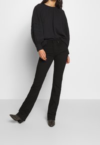 Svart långärmad blus med samlad hals och vertikala veck; tillsammans med svarta utsvängda jeans och mönstrade klackskor.