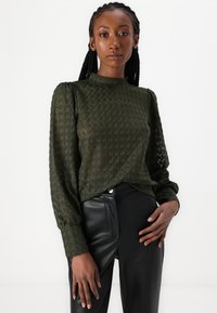 Tamsiai žalia, struktūruota bluzka su aukštu kaklu ir pūstomis ilgomis rankovėmis, derinama su prilipusiais juodais odiniais keliais.