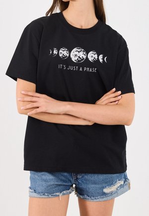 Frau mit verschränkten Armen, die ein schwarzes T-Shirt mit Mondphasen und dem Text "It's just a phase" trägt, kombiniert mit zerrissenen Jeansshorts.