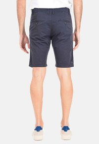 Pantaloni cargo blu navy con vestibilità sartoriale, caratterizzati da bottoni e tasche posteriori, realizzati in un morbido tessuto misto cotone. Scarpe bianche casual visibili.