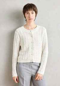 Hvit strikket cardigan med en teksturert mønster, bølget hals og fem knapper foran. Lange ermer og ribbestrikkede mansjetter.