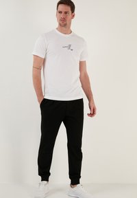White short-sleeve t-shirt with black printed text on the chest, paired with black jogger pants and white sneakers. Simple fit and design.