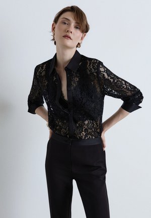 Femme aux cheveux courts portant une chemise noire en dentelle boutonnée et un pantalon noir, posant avec les mains sur les hanches devant un fond uni.