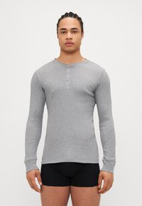 Tricou henley gri cu mâneci lungi, textură ribbed, cu trei butoane la gât și decolteu rotund, purtat cu pantaloni scurți negri strâmți.