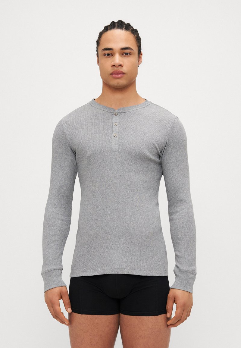 Tricou henley gri cu mâneci lungi, textură ribbed, cu trei butoane la gât și decolteu rotund, purtat cu pantaloni scurți negri strâmți.