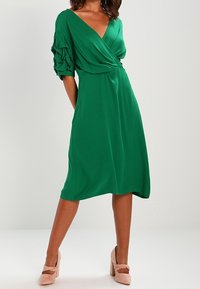 Grön omlottklänning med korta, rynkade ärmar, V-ringning och flödande textur. Avslutad med rosa klackskor med blockhäll.