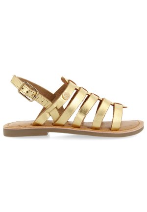 Le sandali metallici dorati presentano un design a fasce con quattro barre che attraversano il piede, una cinghia sul tallone con una fibbia regolabile e una suola flat.