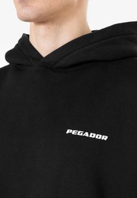 Sudadera negra de tela suave con un bolsillo tipo canguro. Presenta un logo blanco de "PEGADOR" en el pecho y una capucha con cordón.