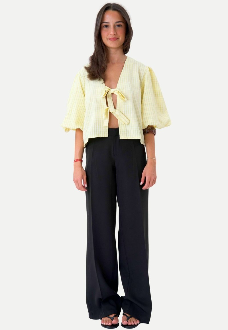 Femme debout portant un chemisier court jaune clair avec des liens sur le devant, un pantalon noir à jambes larges et des sandales noires sur un fond uni.