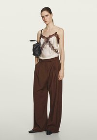 Seidene Cami mit brauner Spitzenborte, kombiniert mit hochgeschnittenen braunen Hosen. Das Model hält eine schwarze geflochtene Tasche und trägt minimalen Schmuck.