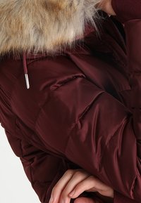 Vinröd quiltad vinterjacka med pälsfoder på huvan och synlig dragsko, bärs av en person som står med armarna korsade.