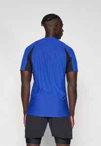 Modra športna majica z teksturiranim dizajnom in črnimi poudarki na ramenih. Kratki rokavi, zaobljen rob in zračen material. Vidne črne kratke hlače.