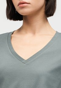 Νεαρό άτομο με κοντά σκούρα μαλλιά που φοράει μια σβηστή πράσινη μπλούζα με V-λαιμόκοψη, απεικονίζεται από το λαιμό μέχρι το πάνω μέρος του στήθους σε απλό φόντο.