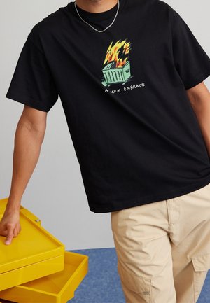 Mand iført sort T-shirt med flaming skraldespands grafik og teksten "A Warm Embrace," beige cargobukser og sølvkæde, lænende på et gult bakke bord.