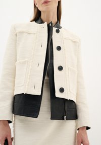 Giacca bianca e testurizzata con fondo in pelle nera. Presenta grandi bottoni neri, due tasche frontali e una chiusura con zip.