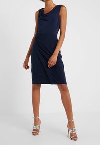 Vestido sin mangas en azul marino con escote drapeado y detalle de lado fruncido. Combinado con sandalias de tacón alto de tiras plateadas. Textura de tela suave.