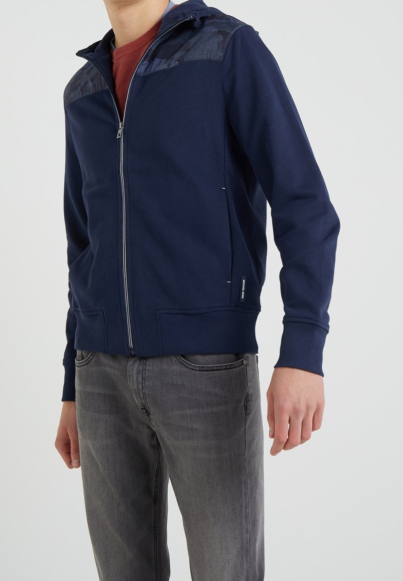 Homme portant un sweat à capuche bleu marine zippé avec des panneaux à motifs aux épaules et un jean gris sur fond uni.