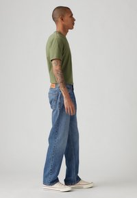 Grøn kortærmet t-shirt parret med lyseblå straight-leg jeans og hvide sneakers. Har tatoverede arme og en afslappet pasform.