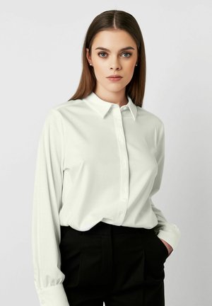 Weißes Langarmhemd mit Button-Down-Kragen und weicher Textur, kombiniert mit schwarzen Hosen. Das Hemd hat eine lockere Passform und einen subtilen Glanz.