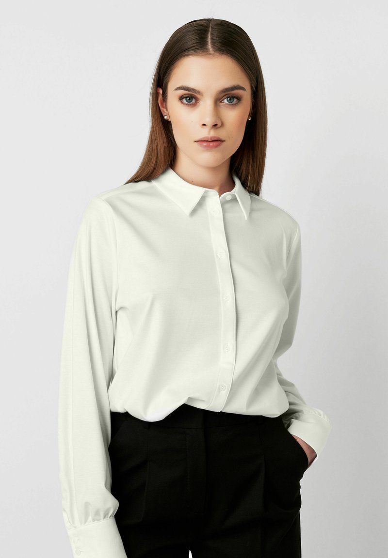 Weißes Langarmhemd mit Button-Down-Kragen und weicher Textur, kombiniert mit schwarzen Hosen. Das Hemd hat eine lockere Passform und einen subtilen Glanz.