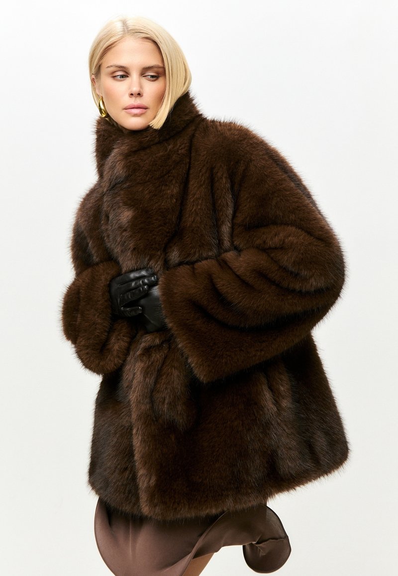 Brun faux fur frakke med høj krave, oversized ærmer og en luksuriøs tekstur. Har en løs pasform og er stylet med sorte handsker.