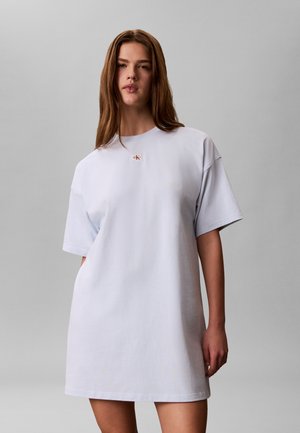 Ung kvinne med langt brunt hår iført en enkel oversized hvit t-skjortekjole med liten Calvin Klein-logo på brystet.