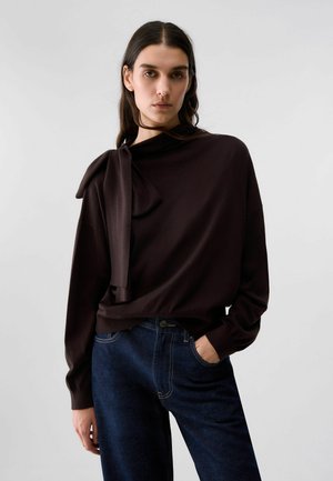 Giovane con lunghi capelli castani indossa una maglia marrone scuro a manica lunga con dettaglio annodato e jeans blu, in piedi davanti a uno sfondo chiaro e semplice.