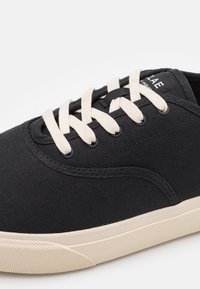 Černé plátěné tenisky s světle béžovými tkaničkami, kulatou špičkou a strukturovanou gumovou podrážkou s jemnými vzory. Značka viditelná na vršku.