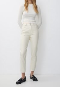 Femme portant un pantalon droit crème taille haute, un haut blanc à manches longues côtelé et des mocassins en cuir noirs, debout contre un mur blanc.