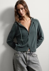 Felpa con zip di colore verde scuro, dal fit rilassato, con polsini e orlo a coste, abbinata a jeans grigi chiari e morbidi. Tessuto liscio, stile casual.