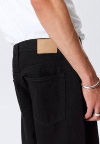 Sorte denimbukser med synligt syningsdesign, to baglommer og et lysebrunt lædermærke med teksten "WEEKDAY." Tekstureret stof med diskret glans.