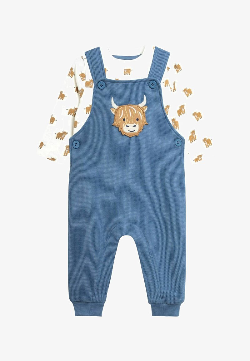 Zils korduroja kombinezons ar pogu aizdari, uz kura krūtīm ir izšūts augstkalnu govs motīvs. Zem tā ir balta krekls ar brūnām govs rakstām.