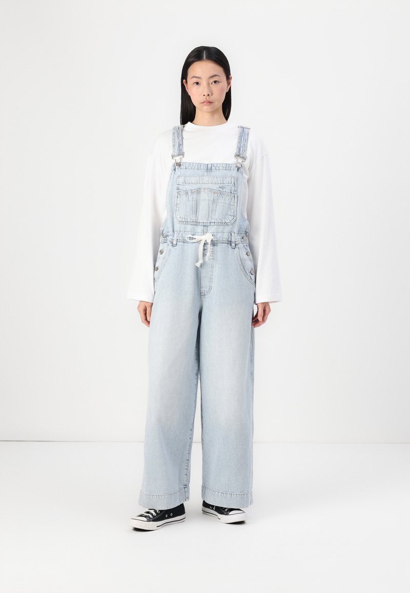 Salopette en denim bleu clair avec une coupe ample, des bretelles réglables, une poche avant, des boutons sur les côtés, et associée à des baskets noires.