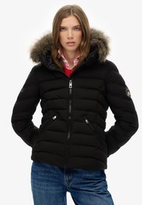 Chaqueta acolchada negra con capucha forrada de piel, cremalleras plateadas, diseño acolchado y bolsillos laterales, combinada con una camisa rosa debajo.