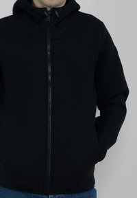 Felpa nera con cappuccio realizzata in un tessuto morbido e texturizzato. Presenta una chiusura a zip completa, due tasche laterali e un cappuccio con coulisse regolabile.