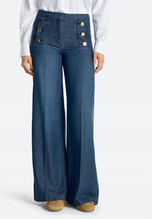 Weite blaue Jeanshosen mit hohem Bund und zwei vertikalen Reihen goldener Knöpfe, getragen mit weißem Hemd und braunen Schuhen.