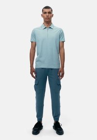 Lyseblå polo med glidelås på kragen, kombinert med tettsittende blå cargo-bukser med sidelommer og elastiske mansjetter. Svarte sneakers.