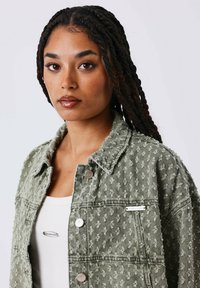 Groene spijkerjasje met een gestructureerd patroon, zilverkleurige hardware accenten en een merketiket. Gecombineerd met een effen wit topje.