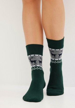 Calze verde scuro con motivi di renne grigi e neri indossate su gambe nude su uno sfondo bianco semplice.