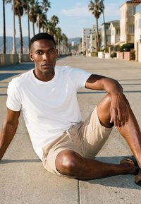 Tânăr în tricou alb și pantaloni scurți bej, stând pe un trotuar însorit, marginat de palmieri și case.