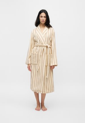 Femme debout pieds nus portant un peignoir long crème avec des rayures verticales marron et deux poches avant, noué à la taille avec une ceinture.