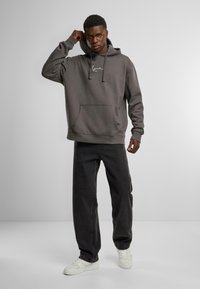 Grå hettegenser med frontlomme og hvit logo, kombineres med mørke jeans og hvite joggesko. Tøyet ser ut til å være strukturert og mykt.