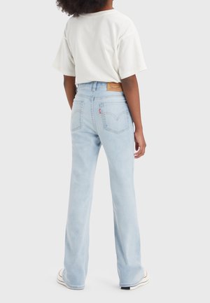 Persoon in lichtblauwe hoge taille jeans en een wit T-shirt, staand met de rug naar de kijker tegen een effen lichte achtergrond.