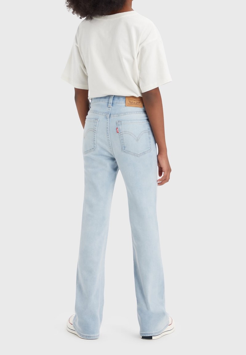 Osoba v světle modrých vysokých džínách a bílém tričku, stojící zády k nám na jednobarevném světle pozadí.