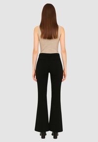 Pantaloni a zampa neri con una texture liscia, vita alta e due tasche posteriori, abbinati a un top beige senza maniche. Vista dal retro.