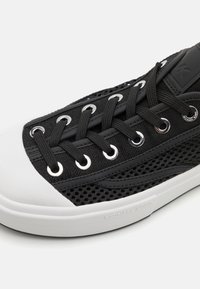 Svart netting-sneaker med hvit gummi tåhette og såle. Har metalløyer og lisser, med en logodetalj på siden.