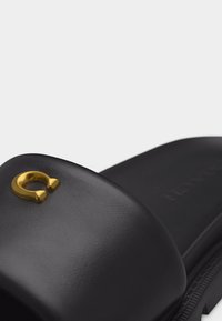 Sandale à glissière noire avec une large bride ornée d'un embellissement doré en forme de fer à cheval sur le dessus.