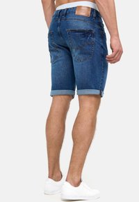 Shorts in denim di un blu scuro, con orli arrotolati, due tasche posteriori e una toppa in pelle in vita. Le sneakers bianche completano il look.
