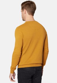 Musztardowy żółty sweter dziewiarski z prążkowanymi mankietami i dołem, z okrągłym dekoltem i gładką fakturą, noszony z ciemnymi dżinsami.